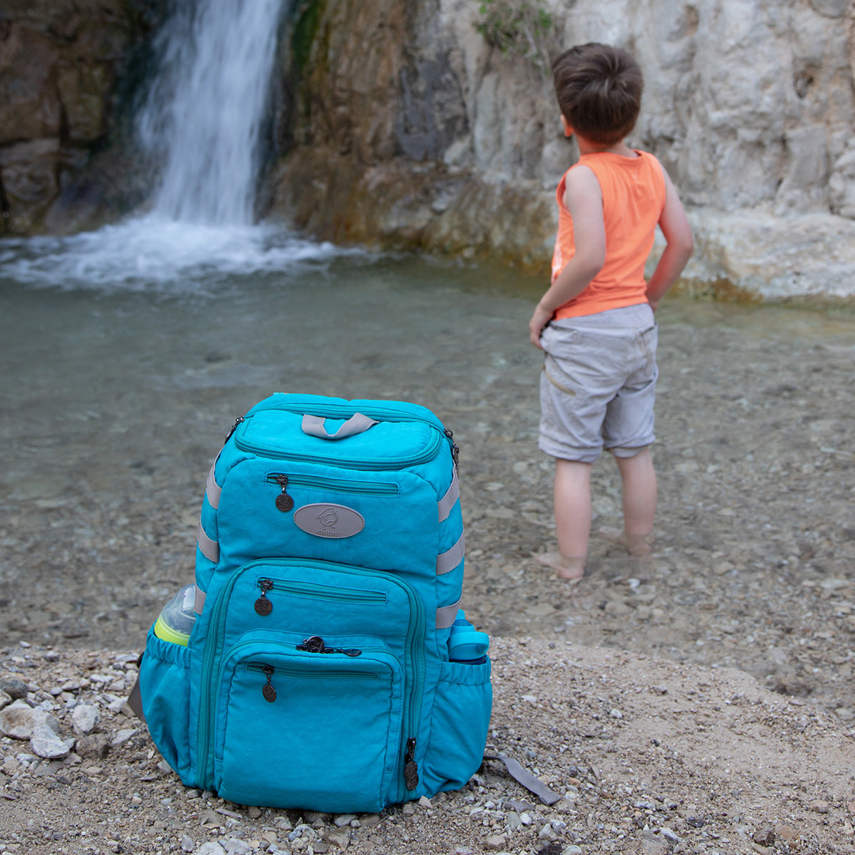 A boy wades into a swimming hole, while his Fledgling bag sits on the shore with dry clothes ready.